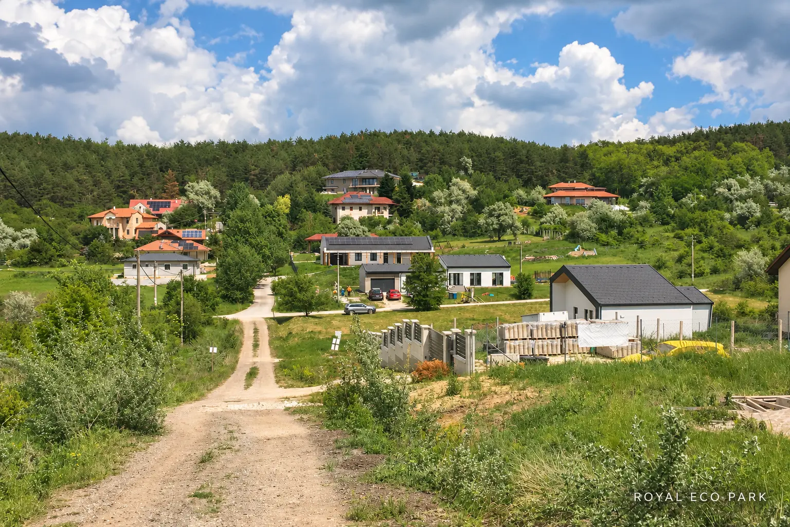 Földút és szóródó házak zöld domboldalon, építőanyagokkal